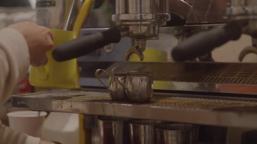 A hand holding a large yellow mug and a metal pitcher are positioned under the nozzles of a professional espresso machine on a counter