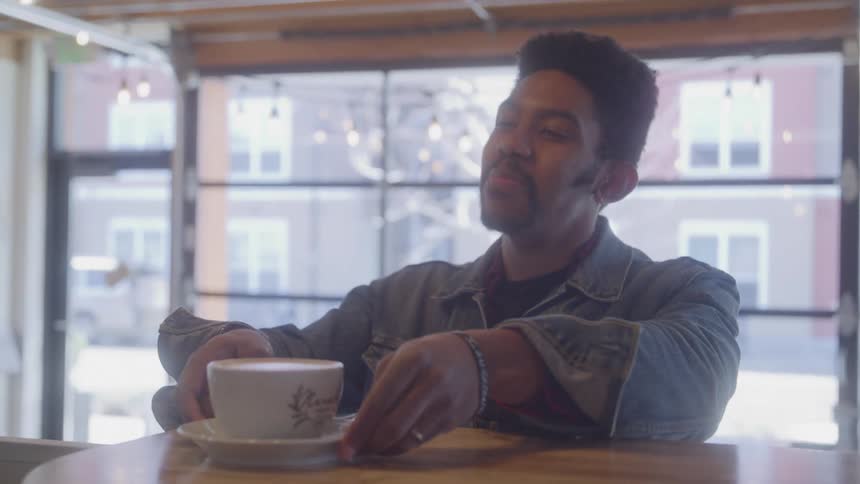 A coffee shop barista hands a cup of coffee to a smiling male customer who then takes the drink and looks away with large glass windows visible in the background
