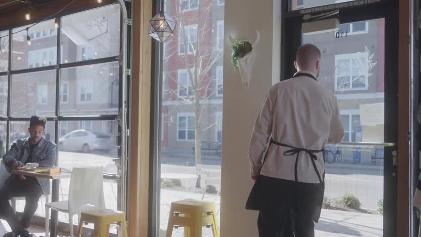 The video shows an employee in a cafe turning a door sign from open to closed and then walking away from the door a customer can be seen sitting at a table near the window