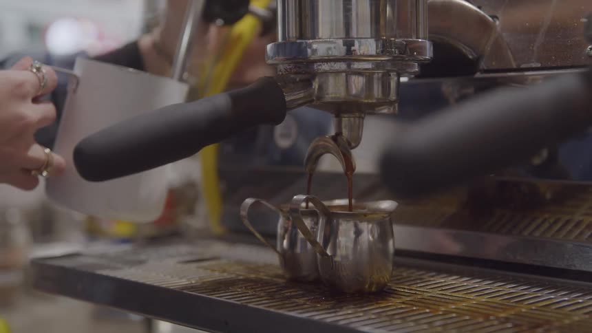 An espresso machine steams milk in a metal pot and coffee drips from a spout into two small pots below