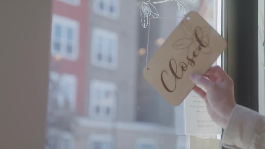 A persons hand turns a sign that hangs in a glass door from showing closed to showing a blank side
