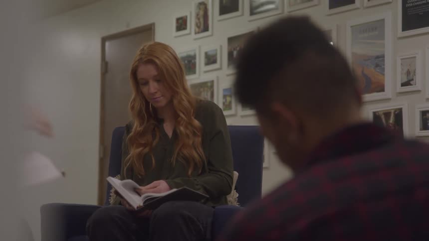A woman reads quietly in a cozy artfilled room while another person sits blurred in the foreground