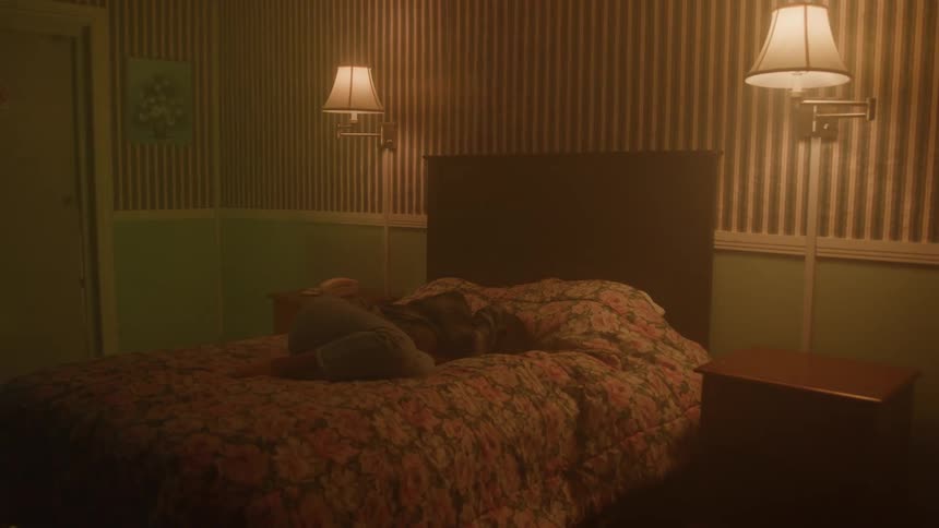 A person lies curled up on a floral bed in a dimly lit bedroom