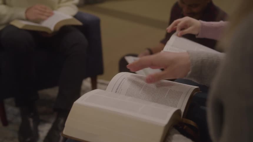 A person flips book pages with their hand while reading aloud from an open book in a group setting