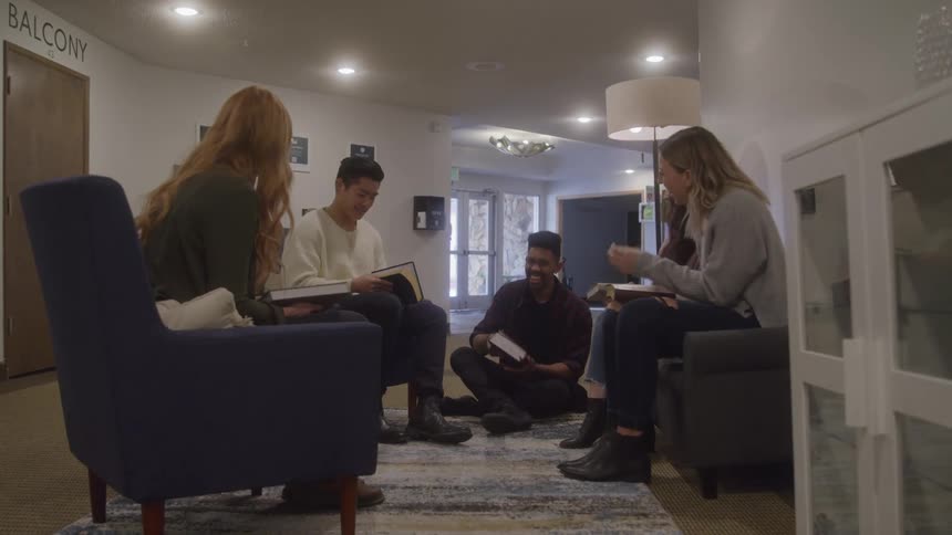 A group of five friends including a man sitting on the floor and four other people in two armchairs are sitting in a circle and talking while holding notebooks and books in a well lit room