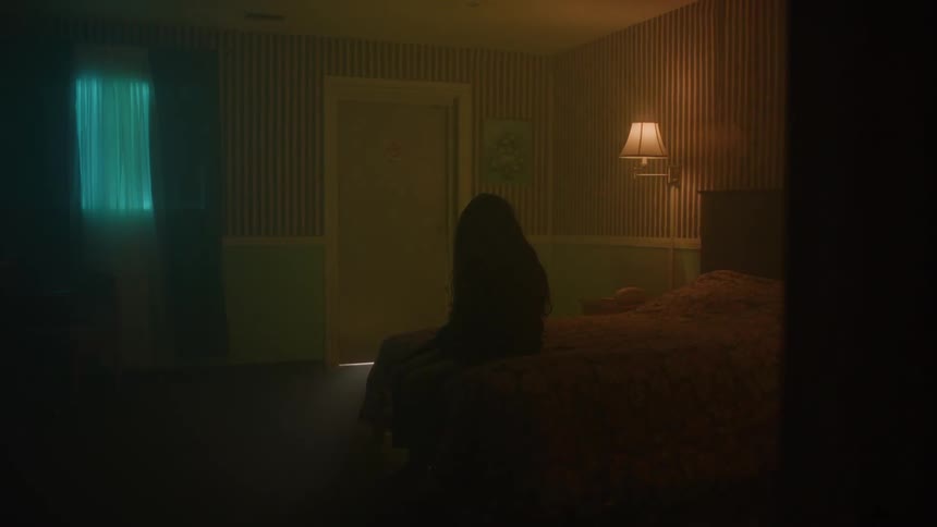 Someone sits on a bed in a dim room lit by a cool window glow