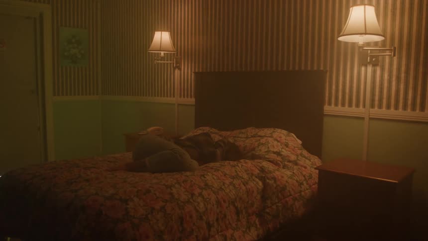 A bedroom with a bed with a person lying on it with hands over their head and theres lamps on the wall on both sides