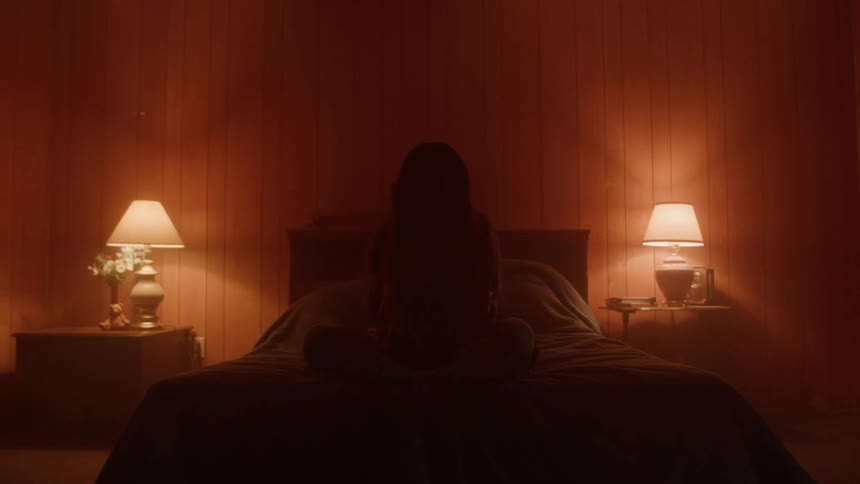The video shows a person with long dark hair sitting on a bed in a dimly lit room with reddishcolored walls and two lit lamps on the bedside tables the person is seen from behind with their head bowed