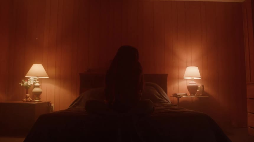 A person sits crosslegged on a bed in a dim warmly lit room
