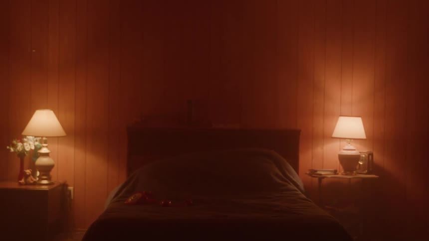 The video shows a person with long dark hair standing in a dimly lit room with reddish walls the individuals back is to the camera and they are standing near the foot of a bed