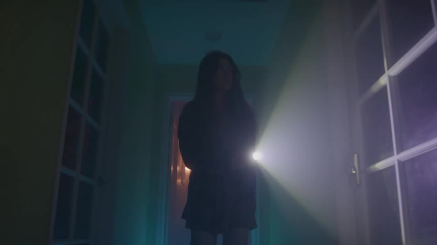 A young woman searches with a flashlight while walking through a dark corridor indoors at night