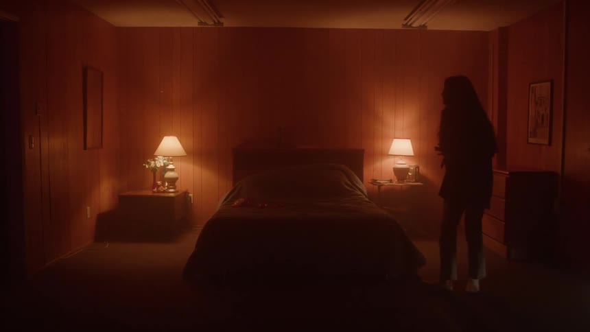 A woman walks past a neatly made bed in a dimly lit wooden room with two lamps casting a warm glow