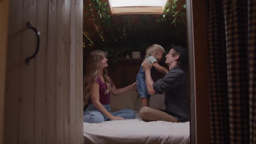 A man and a woman sit on a bed with a child and the man lifts the child up to kiss the child