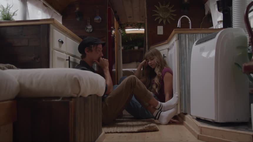 A man and a woman are sitting on the floor of a cozy van interior leaning quietly against the cabinets with thoughtful subdued and somewhat somber expressions