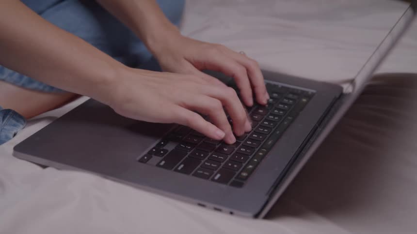The video is a closeup shot of a persons hands as they type on a laptop keyboard they are sitting down and the computer is resting on what appears to be a bed or couch