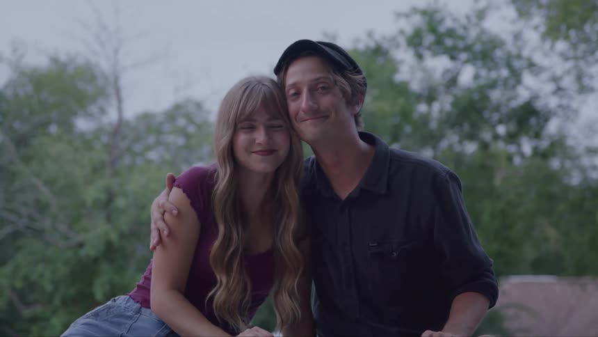 A smiling couple is seated side by side with the woman in a burgundy top and the man in a dark shirt and a black hat as the man puts his arm around the woman and she leans her head against his