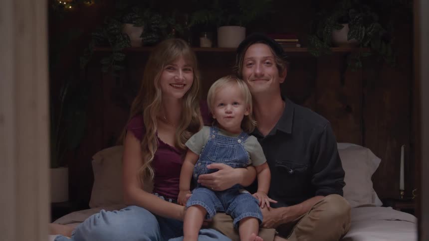 A smiling family sits together on a cozy bed posing for the camera