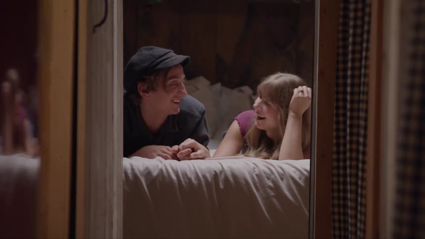 The video shows a man and a woman lying on a bed looking at each other and smiling the woman has long blonde hair and is wearing a pink shirt while the man wears a black hat and a black shirt they appear to be in a bedroom with a window on the right s