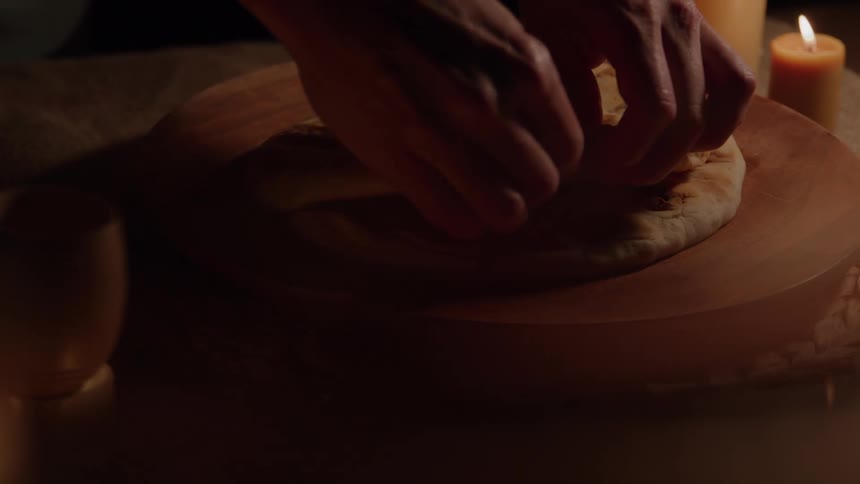 Hands break flatbread on a wooden board beside glowing candles