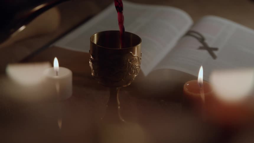 A hand pours liquid into a chalice beside candles and an open book