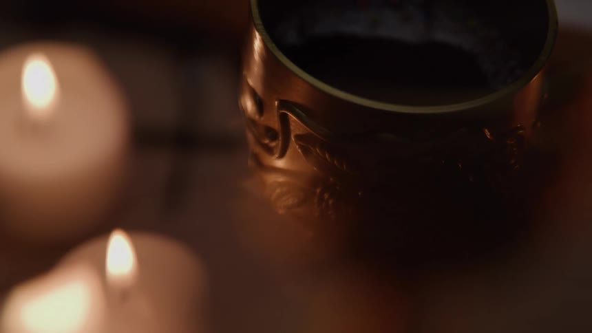 Two softly glowing candles sit beside a reflective cup filled with a dark liquid