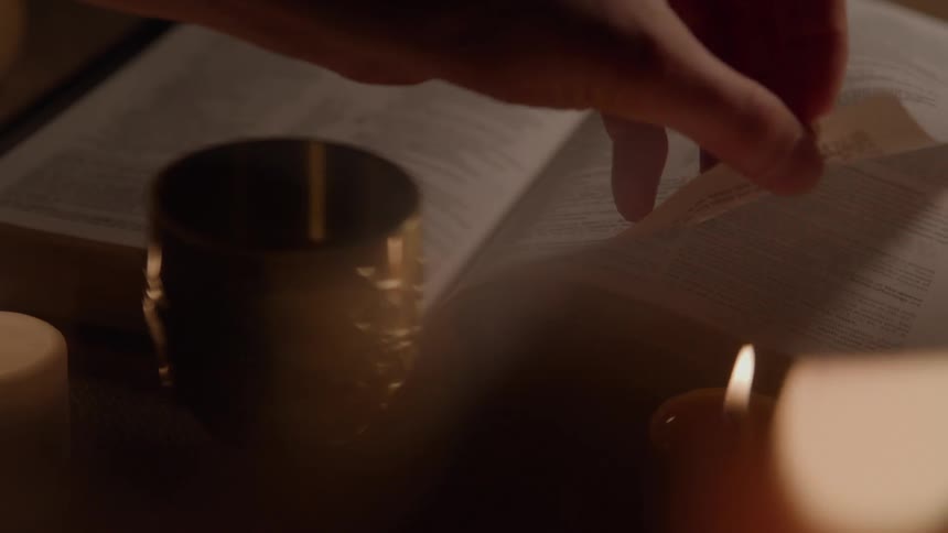 A hand turns a book page beside a candle and metal cup