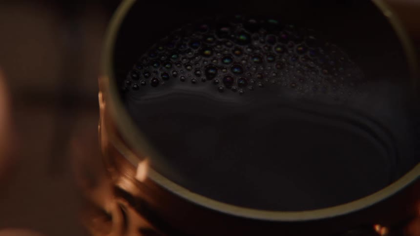 The video shows a closeup shot of a steaming drink in a mug bubbles are visible on the surface of the drink and a blurred candle is in the background