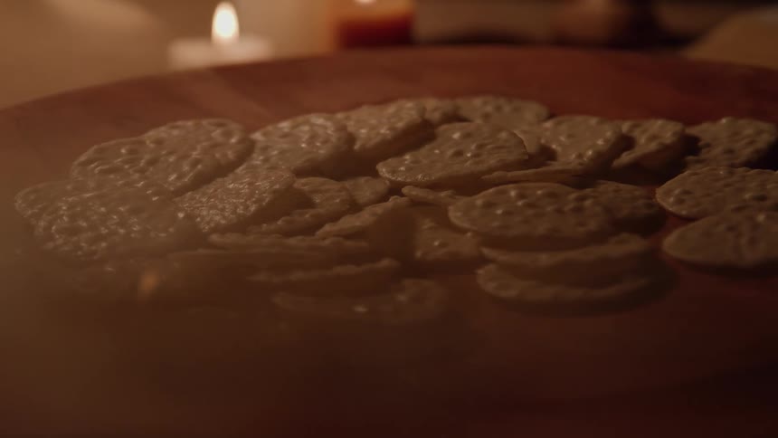 A wooden table holds a pile of thin cookies lit warmly by candlelight
