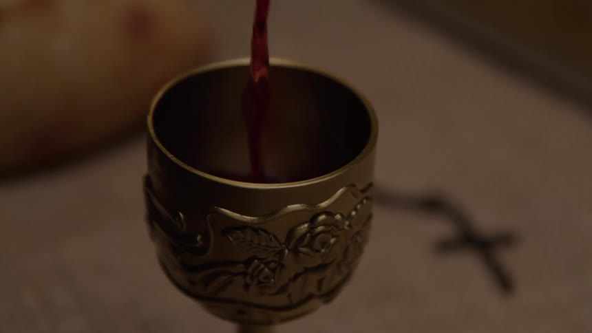 Red wine streams into an ornate chalice on a linencovered table with bread and a cross softly blurred in the background