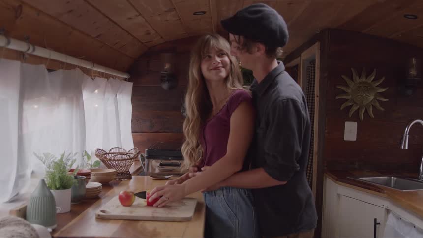 A man walks into a kitchen and affectionately hugs a woman