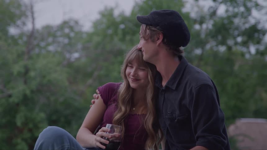 The video shows a man and a woman sitting on a roof or patio looking out at the scenery they are laughing and talking and the man has his arm around the womans shoulder