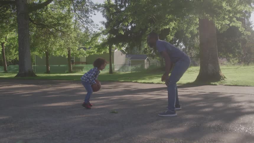 A father and son are playing basketball on a paved court with the father dribbling the ball and the son trying to grab it in a park surrounded by green grass and trees