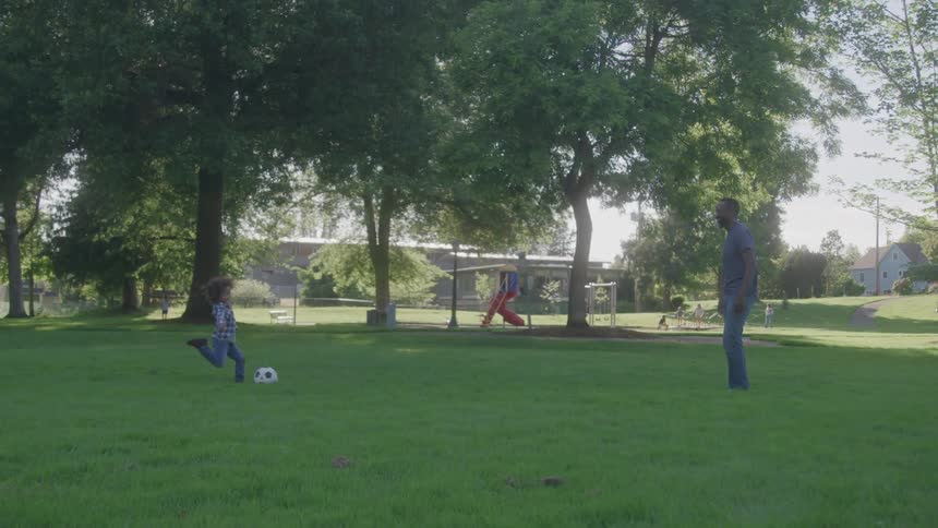 A father and son play soccer in the park on a sunny day