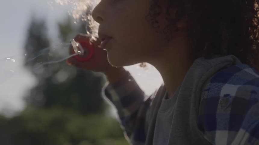 A child exhales to form a delicate bubble from a wand sunlight rimlighting their profile as they play in a green park