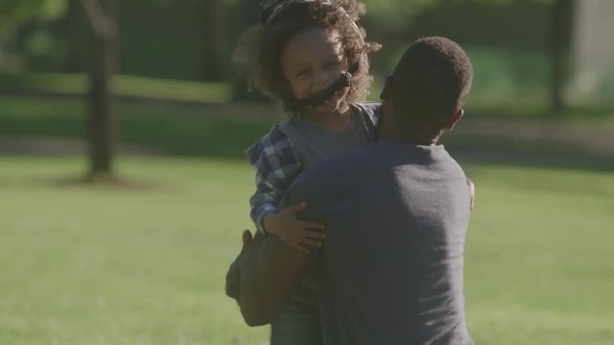 A child wearing a helmet runs on the grass and is hugged by an adult who then falls to the ground