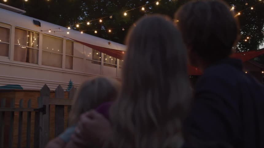 At dusk a small family gazes up at glowing string lights beside their converted bus home wrapped together in an easy contented embrace