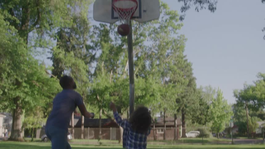 A man and a young child play basketball together outdoors on a sunny day