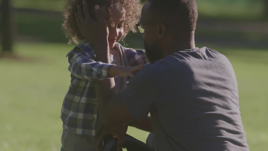 The video shows a man with a beard wearing glasses kneeling down to help a young child with curly hair put on a bike helmet the scene takes place outdoors in a grassy park area on a sunny day