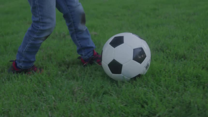 A child runs across a grassy field while casually kicking a soccer ball