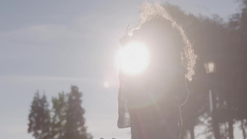 The video shows a child with long curly hair in profile they are standing outdoors silhouetted against a bright hazy sun and are blowing bubbles with a wand