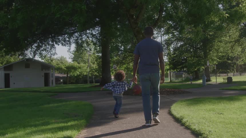 A father and child play basketball in the park on a sunny day