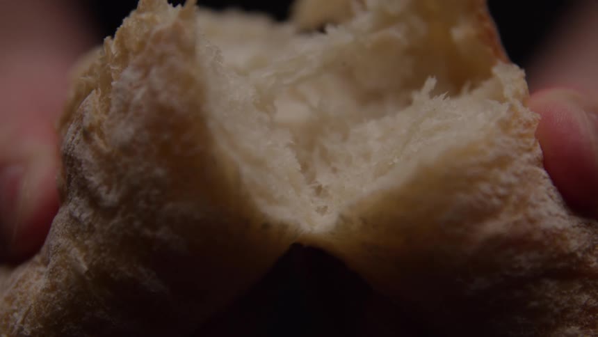A close view shows a persons hands holding a piece of crusty brown bread with a light layer of flour tearing it in half to reveal the soft white interior