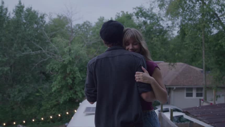 The video shows a man and a woman from the back as they dance together on a rooftop or a porch they are surrounded by lush trees and string lights