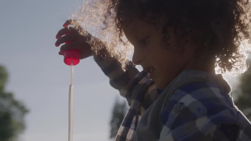 The video shows a child with curly hair in a plaid shirt the child is playing with a bubble wand blowing bubbles into the air with the sun shining behind them