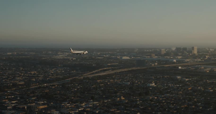 An airplane flies over a city with mountains and a highway in the background