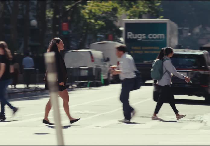 A crowd of people walks on a sidewalk and crosses a street with vehicle traffic including a yellow taxi a grey suv and a red car all passing in front of a storefront and a food stand