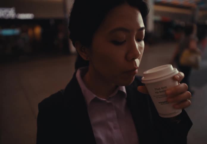 A woman in a blazer and shirt stands in an indoor space holding a paper cup and raising the cup to her mouth to drink from it