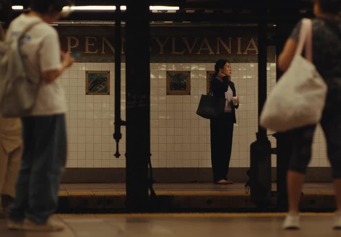 The video shows a woman in a black suit standing alone on a subway platform other people are blurred in the foreground walking on the platform