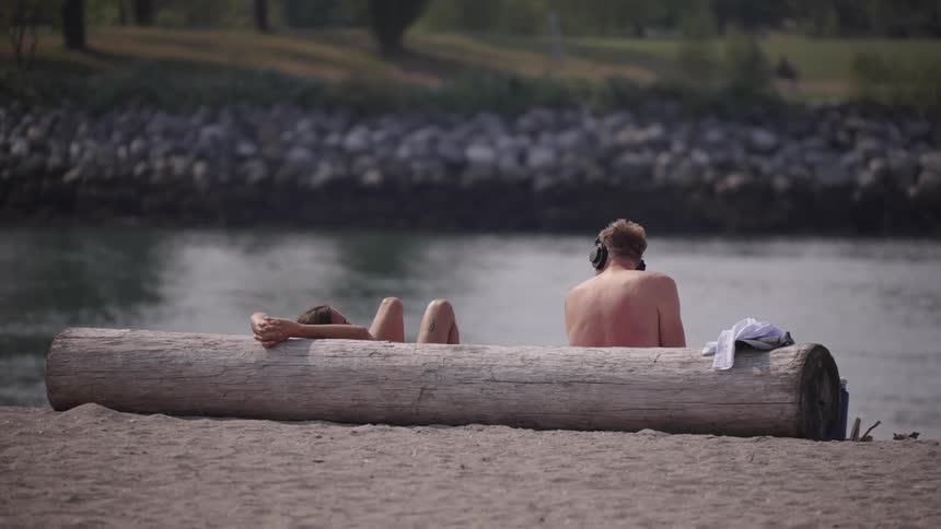 Two shirtless young men relaxing by a river near a fallen tree trunk one of them lying back against it and the other sitting up and wearing large headphones