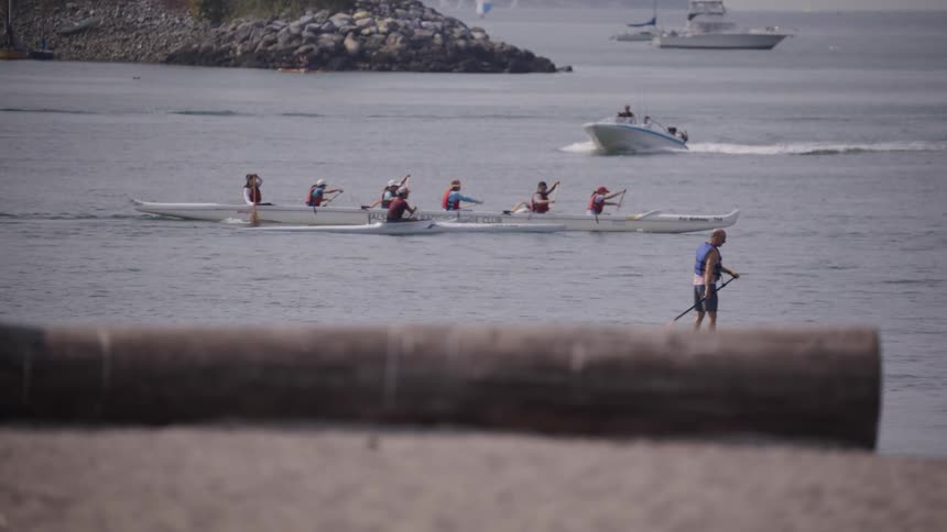 A group of people in an outrigger canoe paddle through a body of water while a person on a paddleboard stands in the foreground and other boats move and sit on the water in the background